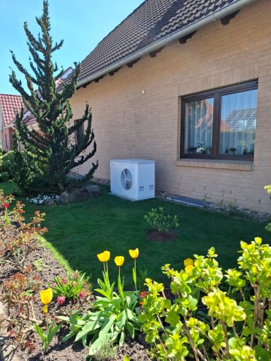 Weiße Luft-Wasser-Wärmepumpe im Gartenbereich direkt neben einem Einfamilienhaus.